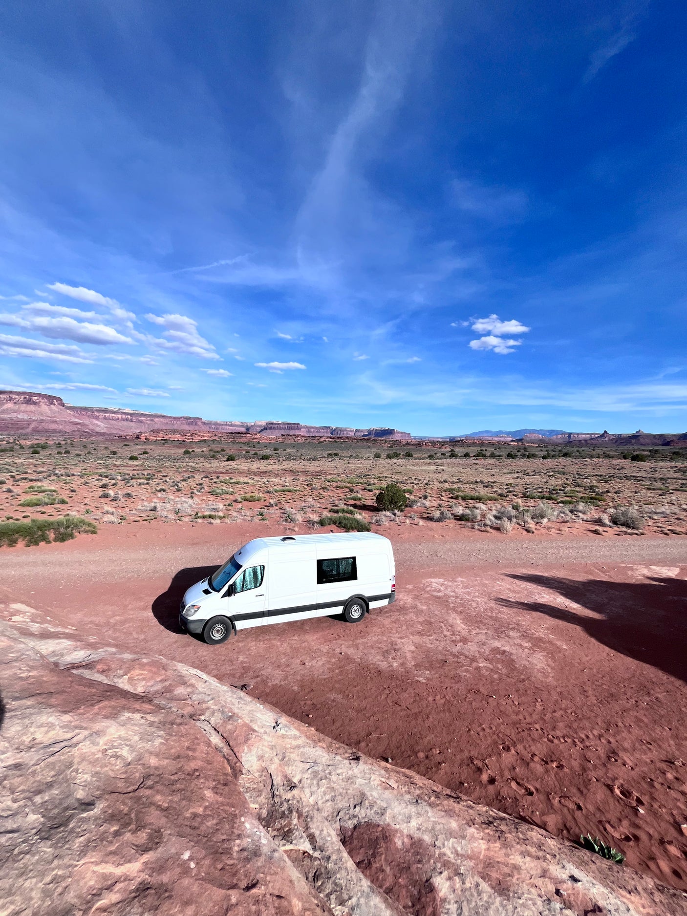 A white RV rental is parked in a vast red desert landscape with distant mesas under a bright blue sky.