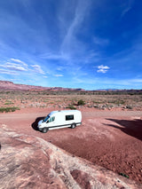 A white RV rental is parked in a vast red desert landscape with distant mesas under a bright blue sky.