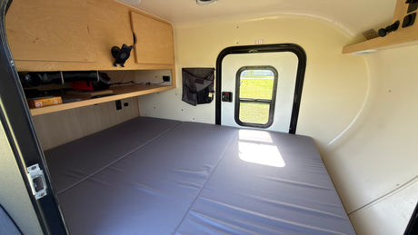 RV rental interior: wide bed, wooden cabinets, and rear exit door with view to outside grass.