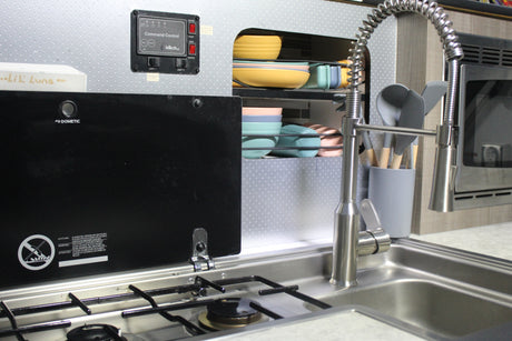 Compact RV rental kitchen with Dometic stove, sink, colorful dishes, and an InTech control panel for water and power.