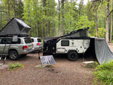 Explore the wilderness! RV rental featuring an SUV with a rooftop tent, overland trailer, and solar power setup in a lush forest.