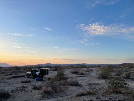 A person prepares camp with an RV rental SUV in the desert at sunset, surrounded by sparse brush & distant mountains.
