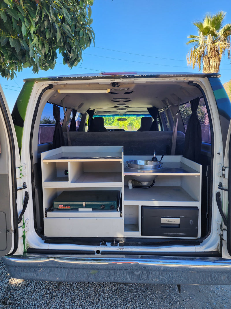 Rear view of an RV rental camper van's custom kitchen: sink, pull-out stove, Dometic fridge, and storage shelves. — photo 5