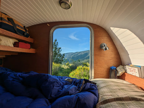 Serene rv rental interior: blue sleeping bag on bed, wood/white panels, with a window view of sunny mountains & yellow flowers.