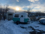 Scenic winter camping with an RV rental: a charming teal and white travel trailer amidst snow, surrounded by other RVs at sunset.