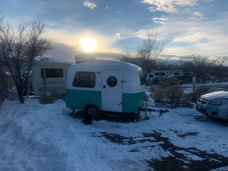 Scenic winter camping with an RV rental: a charming teal and white travel trailer amidst snow, surrounded by other RVs at sunset.