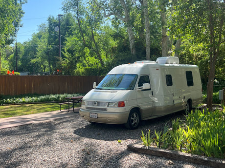 A cream Class B RV parked on a gravel campsite next to a picnic table, perfect for your next rv rental.