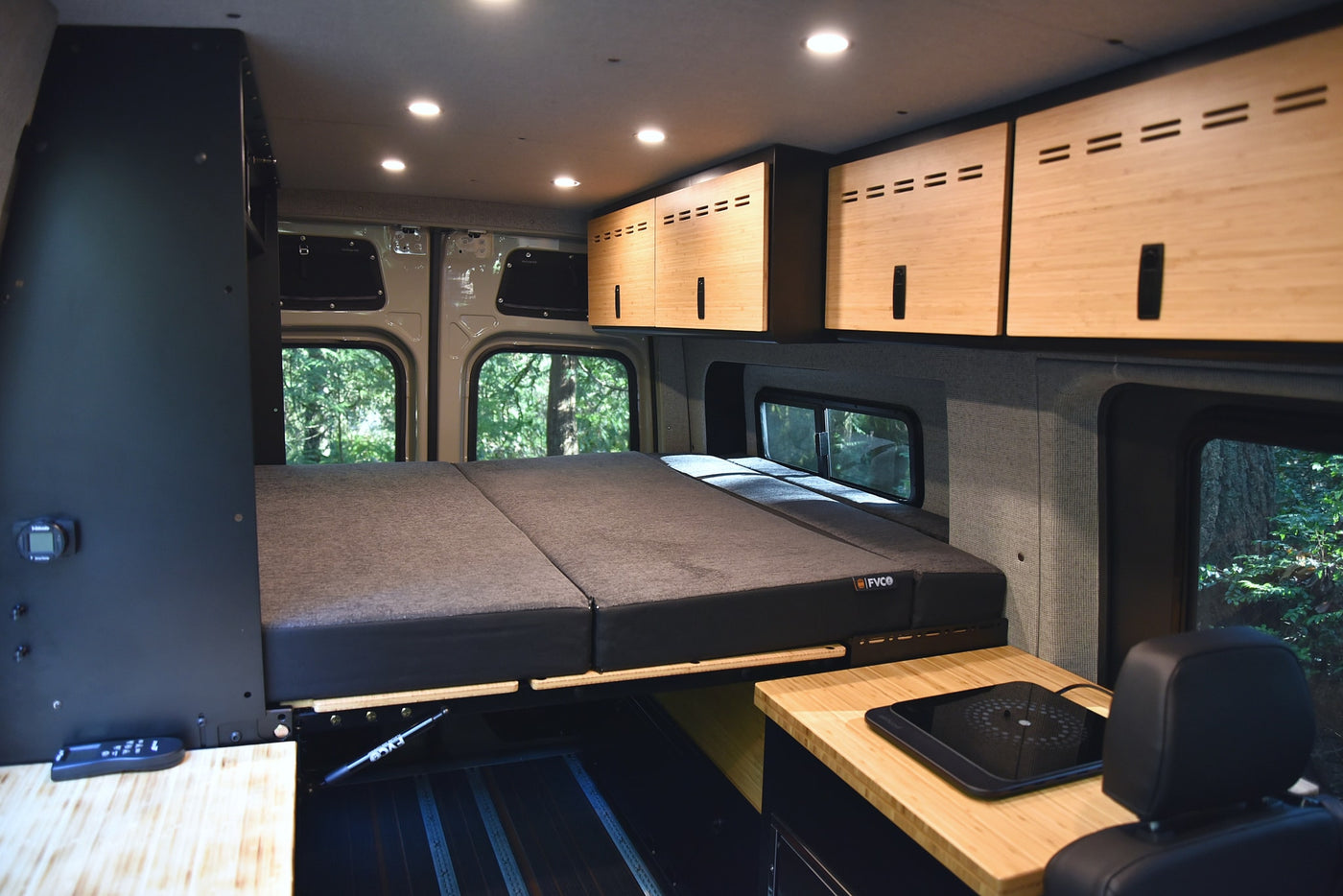 RV rental: interior with a comfortable foldable bed, sleek wood cabinets, and cooktop. Lush nature visible through windows. — photo 8