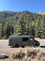 Gray camper van on a mountain road beside pine trees. Your next RV rental adventure awaits here!