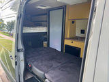 Spacious RV rental interior with bunk bed, convertible lower bed, kitchenette, and fridge, viewed from open side door.