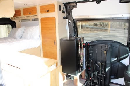 Cozy RV rental interior featuring a bed, wooden storage, fridge, and adaptable seating. Perfect for mobile adventures.