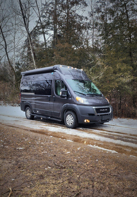 Dark grey camper van RV rental on a snowy, muddy path. Bare and evergreen trees line the winter road.