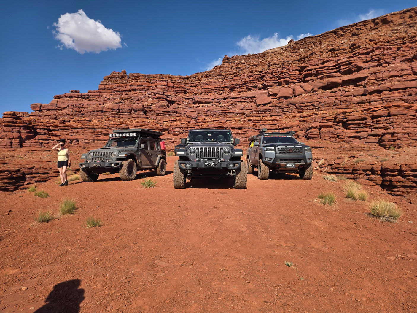 Three off-road vehicles, one with an overlanding setup (like an RV rental), on red desert terrain with rock formations & a woman.