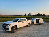 A white pickup truck tows a compact teardrop RV rental camper, parked on gravel beside a lush vineyard at sunset.