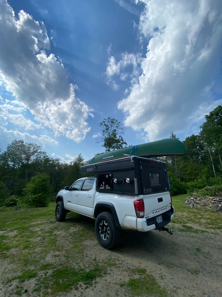 A white Toyota Tacoma truck with a bed camper and canoe is parked outdoors. Perfect RV rental for outdoor adventure!
