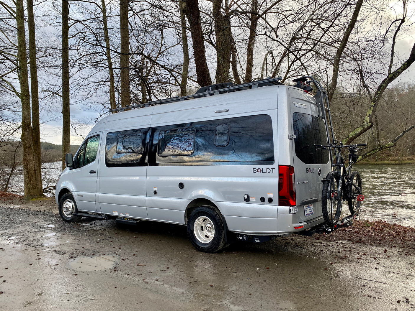 Silver Winnebago Boldt rv rental parked by a muddy river, ready for adventure with bikes on the back.
