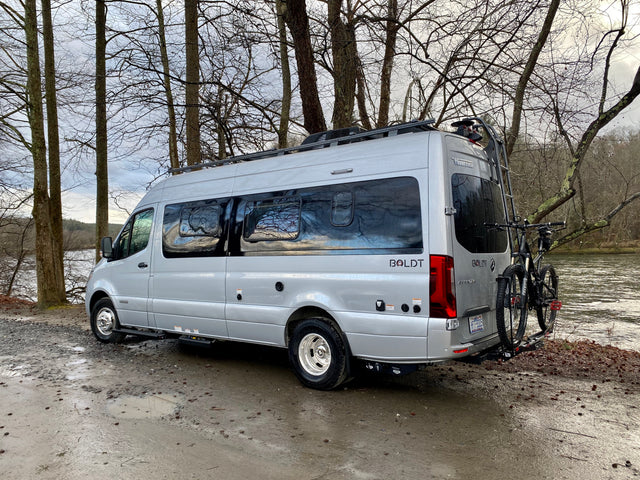 Silver Winnebago Boldt rv rental parked by a muddy river, ready for adventure with bikes on the back.