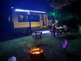 A family enjoys a cozy night camping in their RV rental. A campfire glows as they gather around a picnic table with bright lights.