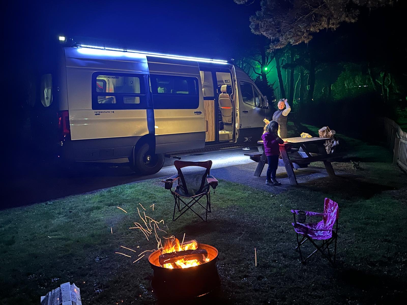 A family enjoys a cozy night camping in their RV rental. A campfire glows as they gather around a picnic table with bright lights.