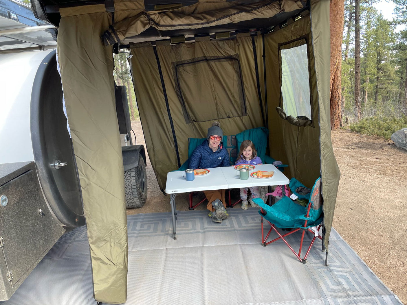 Two people enjoy a meal inside an RV rental tent annex, extending their cozy outdoor living space in the forest.