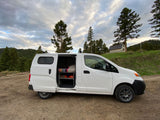White camper van RV rental, ready for adventure! Side door open, revealing bed & storage, on a dirt road by pine trees.