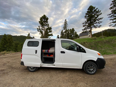 White camper van RV rental, ready for adventure! Side door open, revealing bed & storage, on a dirt road by pine trees.