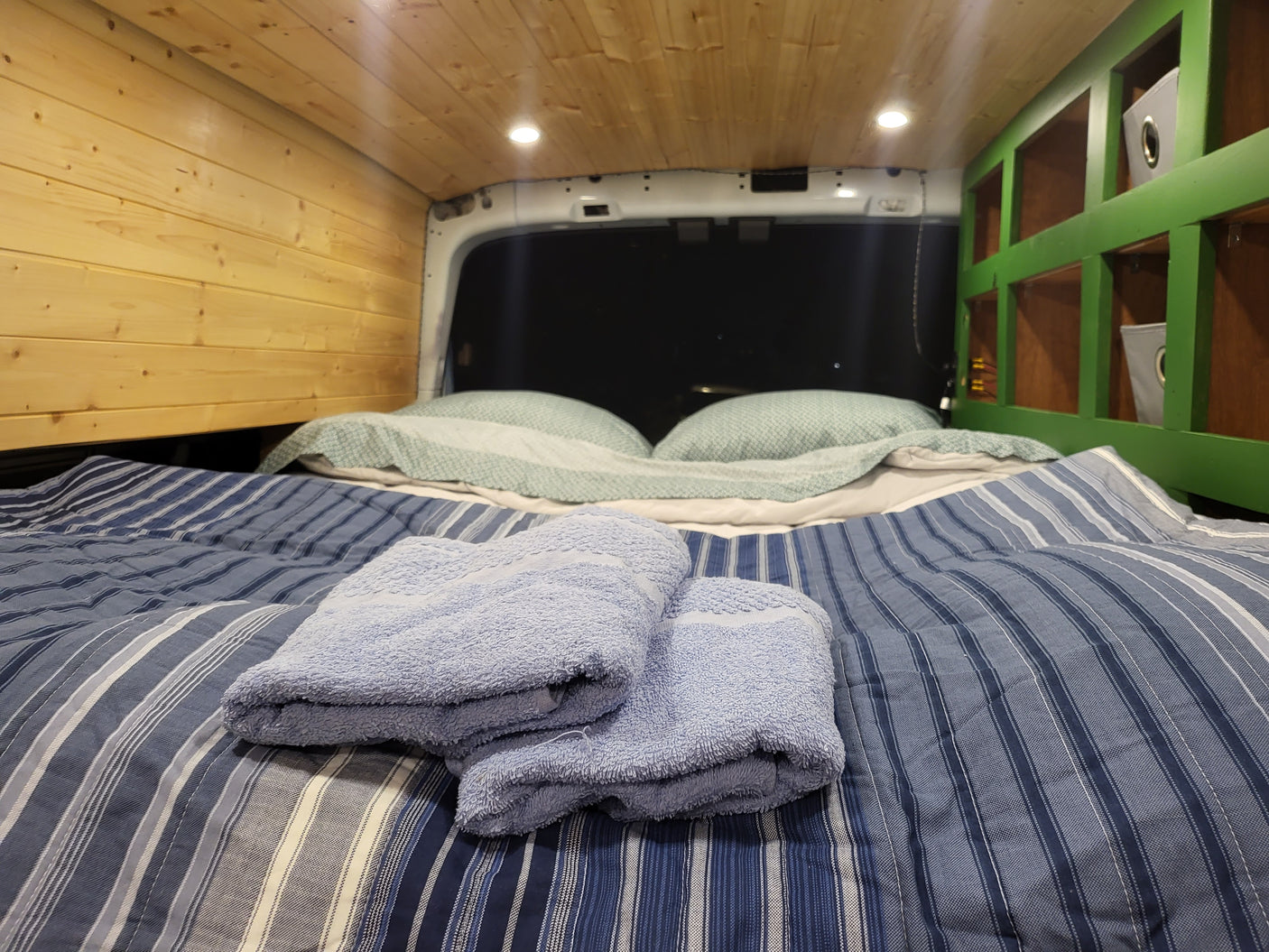Cozy RV rental interior with striped bed, fluffy towels, wood paneling, and green storage shelves. Ready for adventure!