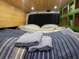 Cozy RV rental interior with striped bed, fluffy towels, wood paneling, and green storage shelves. Ready for adventure!