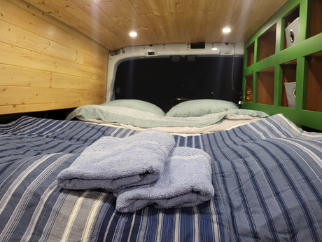 Cozy RV rental interior with striped bed, fluffy towels, wood paneling, and green storage shelves. Ready for adventure!