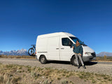 An RV rental van, with bike, and a person stand before the majestic, snow-capped Grand Teton mountains.