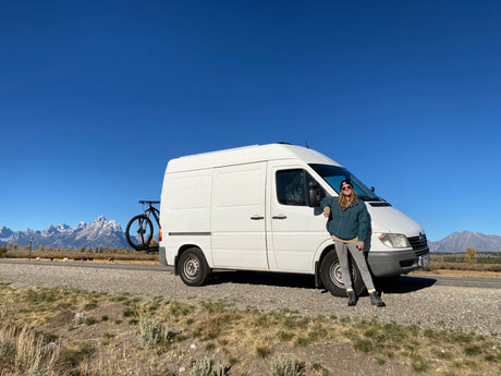 An RV rental van, with bike, and a person stand before the majestic, snow-capped Grand Teton mountains.