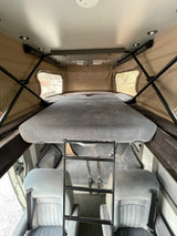 Upper sleeping berth of an RV rental camper van, accessed by a ladder. Features pop-top canvas windows and lower cabin seats.