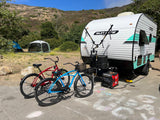 RV rental parked at a sunny mountain campsite with bikes, tents, and a generator on pavement with kids' chalk art.