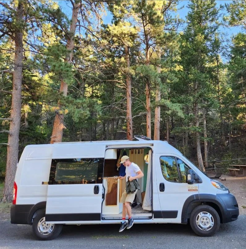 White RV rental camper van parked in a forest. A person steps out from the open side door into the sunny nature.