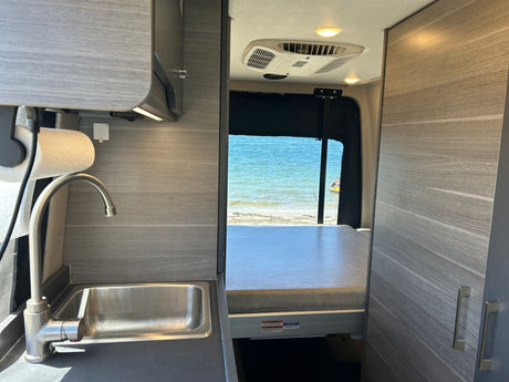 Inside a modern rv rental: a well-equipped kitchen with sink, AC, and a comfortable bed overlooking a serene ocean view.
