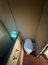 Compact RV rental bathroom: Features a white toilet, trash can, and a toilet paper holder. Wood floor and bright interior.