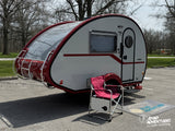 A red and white nuCamp T@B 320 teardrop RV rental, parked with a red folding chair & "Road Adventures" mat for camping.