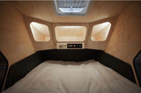 Cozy RV rental interior: bed, overhead cubbies, and a control panel. Compact, light wood paneled camper bedroom with skylight.