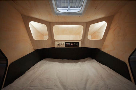 Cozy RV rental interior: bed, overhead cubbies, and a control panel. Compact, light wood paneled camper bedroom with skylight.