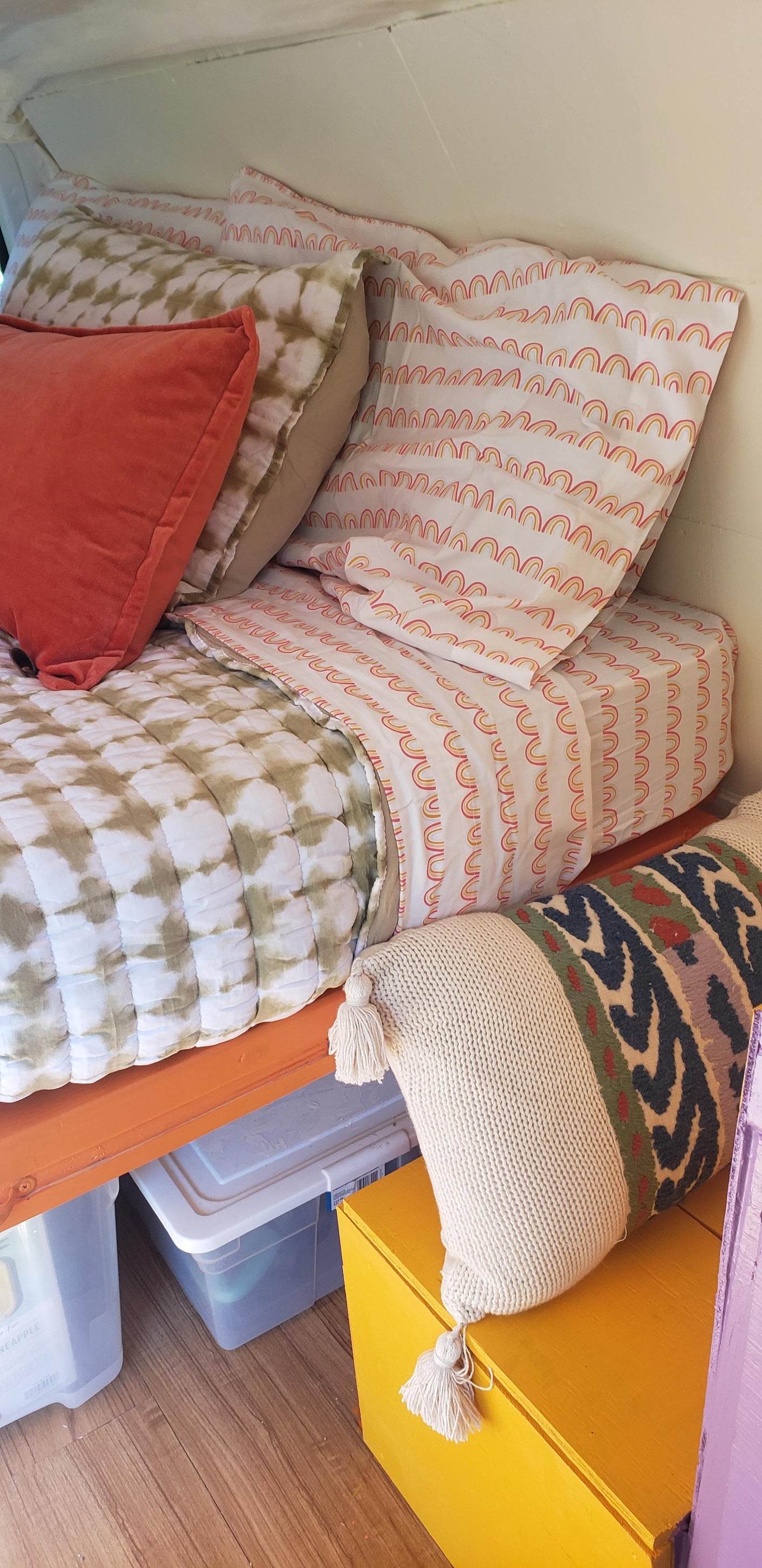 Cozy RV rental bed with colorful rainbow & tie-dye pillows, plus practical yellow storage and under-bed bins.