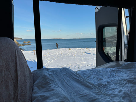 View from an RV rental's open back doors: a person on a snowy beach looks out at the ocean and distant lighthouse.