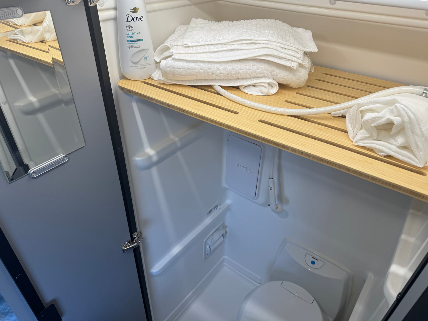 This RV rental's compact wet bath features a toilet, shower, and amenities like towels & Dove body wash on a wooden shelf.