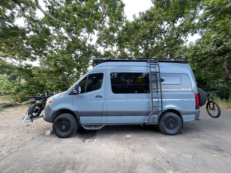 A grey 4x4 Sprinter camper van, equipped with bikes and roof rack, parked outdoors. Great for rv rental adventures.