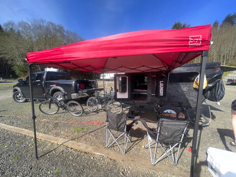 Sunny day camping with an rv rental setup: black truck, compact trailer, bikes, chairs, and a bright red canopy. Outdoor adventure!