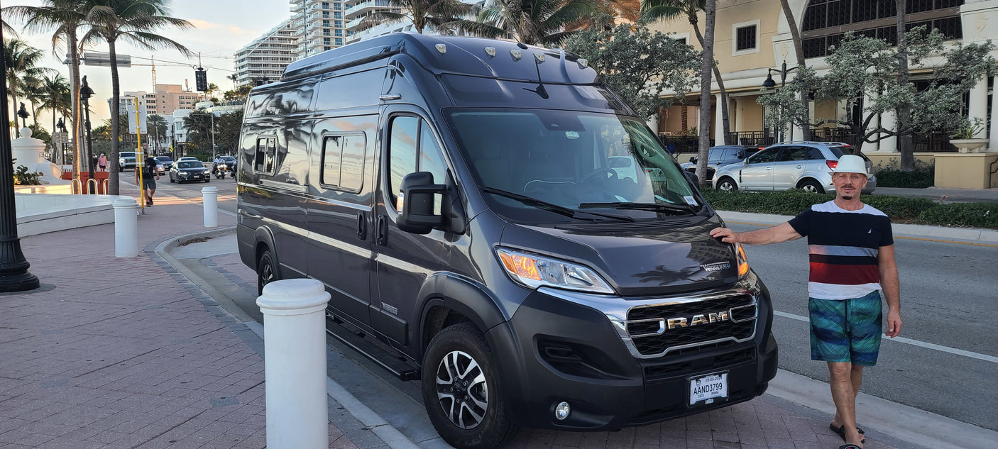 A man poses proudly beside his gray Ram ProMaster RV rental, parked on a palm-lined street in a vibrant city scene.