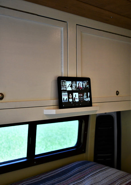RV rental interior: White cabinets above a window, with a tablet displaying Netflix on a shelf. A bed is below.