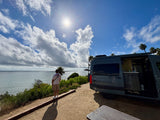 A person views the sparkling ocean from a scenic cliffside spot, with a modern RV rental parked nearby on a bright, cloudy day.