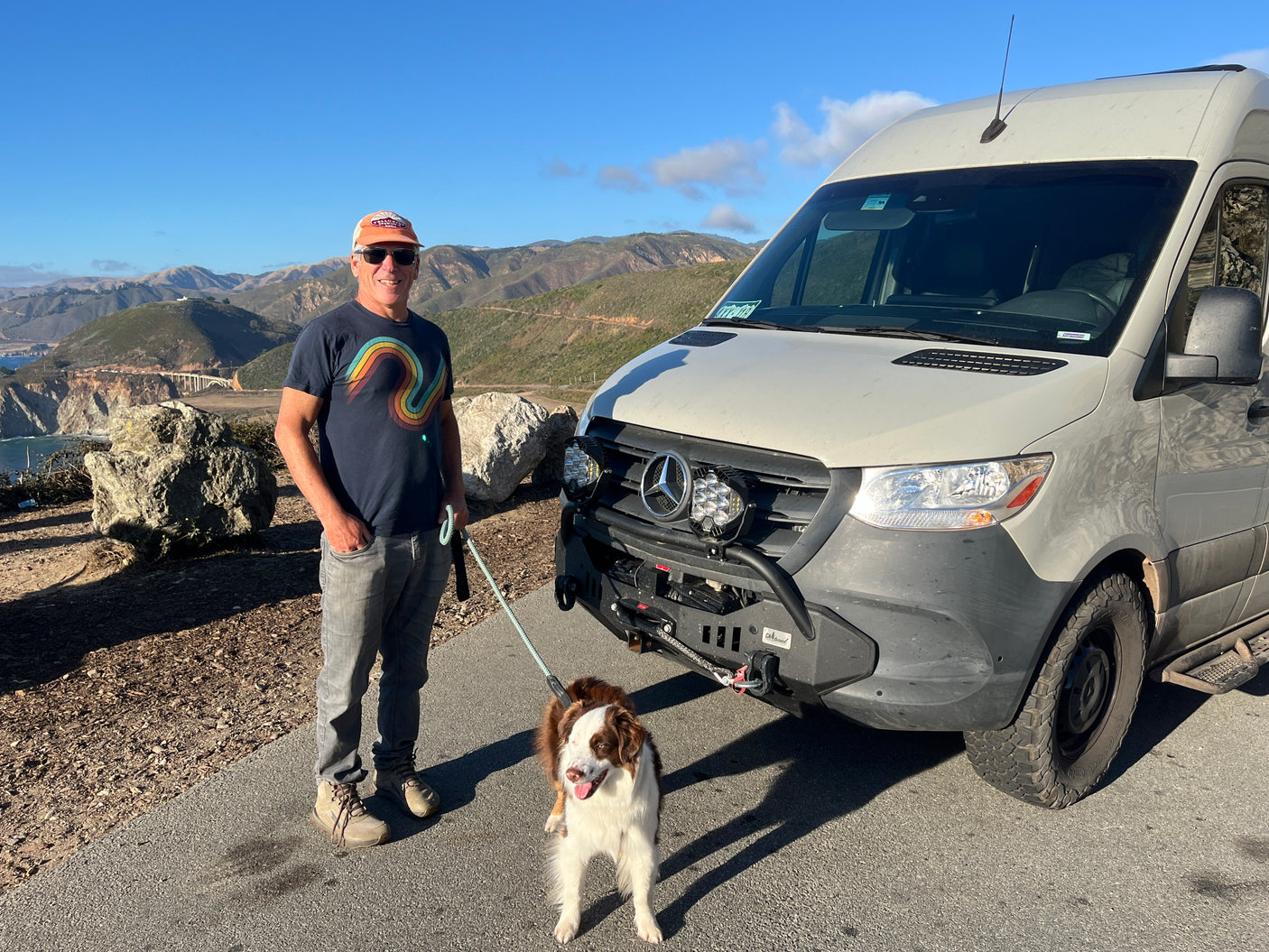 Smiling man and dog with an **RV rental** camper van overlooking scenic coastal mountains and a bridge.