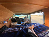 Inside an RV rental, a man relaxes in the driver's seat. Feet are propped on a cozy blanket, enjoying the van's custom interior and the view.