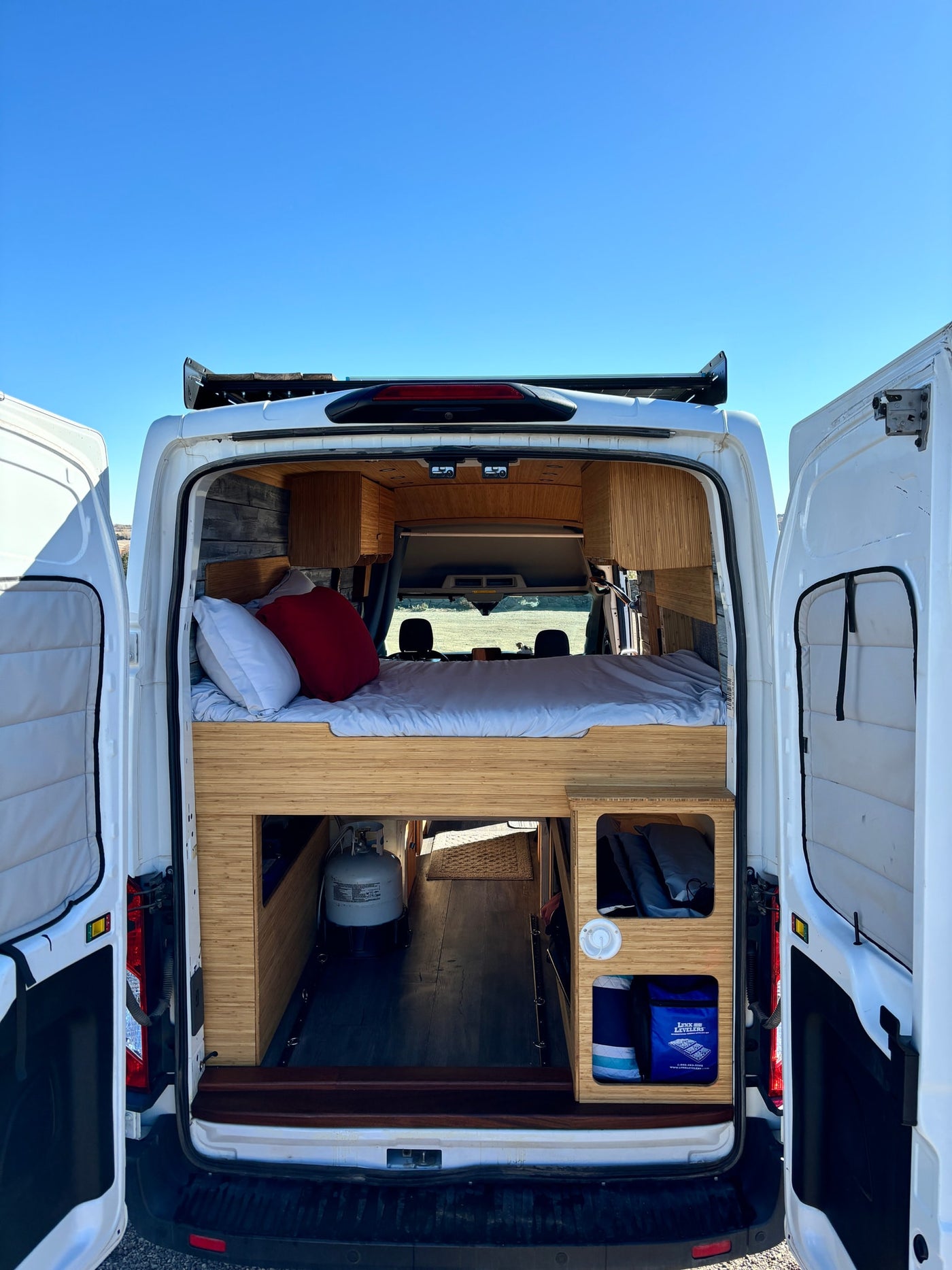 Open doors reveal an RV rental camper van interior: bed, bamboo storage, and propane tank for travel. — photo 7